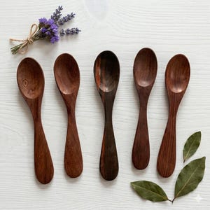 May include: Five wooden spoons with varying shades of brown, arranged in a row. The spoons have long, slender handles and rounded bowls. A bundle of purple flowers and green leaves are placed near the spoons, creating a natural aesthetic.