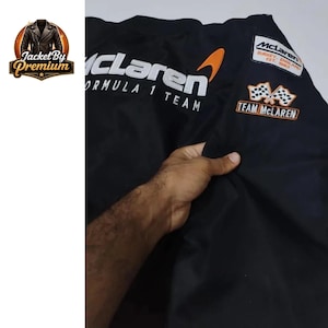 May include: Black McLaren Formula 1 Team jacket with white and orange accents. Features the McLaren logo and text, along with a Team McLaren patch. The jacket has a classic design.