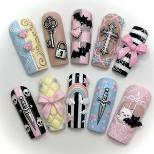 May include: A collection of ten press-on nails with various designs. The nails feature a variety of colors, including pink, black, white, and blue. Designs include bats, keys, crosses, bows, skulls, rainbows, and floral patterns. Each nail has a unique, detailed design.