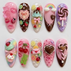 May include: A collection of ten artificial nails with various designs. The nails feature a variety of colorful decorations, including gummy bears, lips, ladybugs, cherries, ice cream cones, rainbows, and lollipops. The nails are painted in shades of pink, brown, and purple.