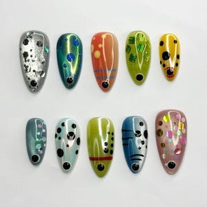 Può includere: Dieci unghie finte a forma di mandorla con design vari, tra cui argento metallizzato con glitter esagonali, verde iridescente con pois blu, arancione a righe, verde lime con foglia, giallo con pois neri e rosa con paillettes colorate.