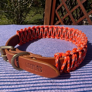 Puede incluir: Un collar de perro de cuero marrón con un diseño de cuerda naranja y blanca tejida. El collar tiene una hebilla de latón y una etiqueta de cuero con el nombre "COOPER'S". El collar está sobre una superficie a rayas azules y blancas.