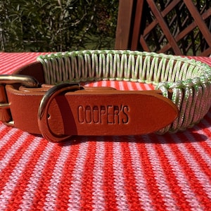 Puede incluir: Un collar de perro de cuero marrón con una hebilla de latón y una sección de nailon verde y blanco tejido. El collar está grabado con el nombre "COOPER'S". El collar está sobre una superficie a rayas rojas y blancas.