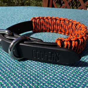 Puede incluir: Collar de perro de cuero negro con una sección tejida de paracord naranja y negro. El collar tiene una hebilla de metal plateado y una etiqueta de cuero negro con el nombre "COOPER'S" en relieve.