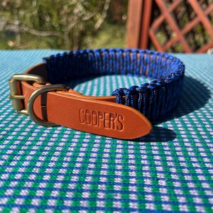 Puede incluir: Un collar para perros trenzado azul y negro con una correa de cuero marrón y una hebilla de latón. La correa de cuero está grabada con el nombre "COOPER'S". El collar está sobre una superficie a cuadros azules y verdes.