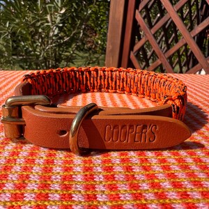 Puede incluir: Un collar para perros con un diseño tejido en naranja y negro, combinado con una correa de cuero marrón y herrajes de latón. La correa de cuero está grabada con el nombre "COOPER'S".