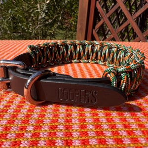 Puede incluir: Un collar de perro de cuero marrón con una sección tejida de paracord naranja, verde y blanco. El collar tiene una hebilla plateada y una anilla en D. El nombre "COOPER'S" está grabado en el cuero.