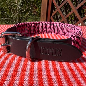 Puede incluir: Un collar de perro de cuero negro con un diseño tejido rosa y negro. El collar tiene una hebilla plateada y una etiqueta con el nombre "COOPER'S" en relieve. El collar está sobre una superficie a rayas rojas y blancas.