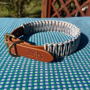 Puede incluir: Un collar de perro de cuero marrón con un diseño tejido en azul y beige. El collar tiene una hebilla de latón y una etiqueta de cuero con la inscripción "COOPER'S". Descansa sobre una tela a cuadros azul y verde.