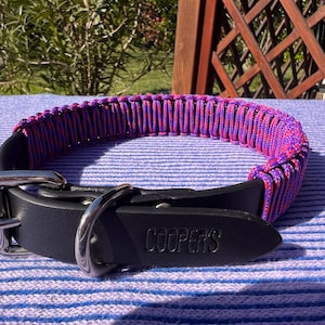 Puede incluir: Un collar de perro de cuero negro con un diseño tejido en morado y rosa. El collar tiene una hebilla plateada y una anilla en D de metal. El nombre "COOPER'S" está grabado en el cuero.