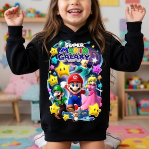 May include: Black hooded sweatshirt featuring a colorful graphic of Mario, Luigi, Princess Peach, and other characters from the Super Mario Galaxy video game. The design includes stars and the game's logo.