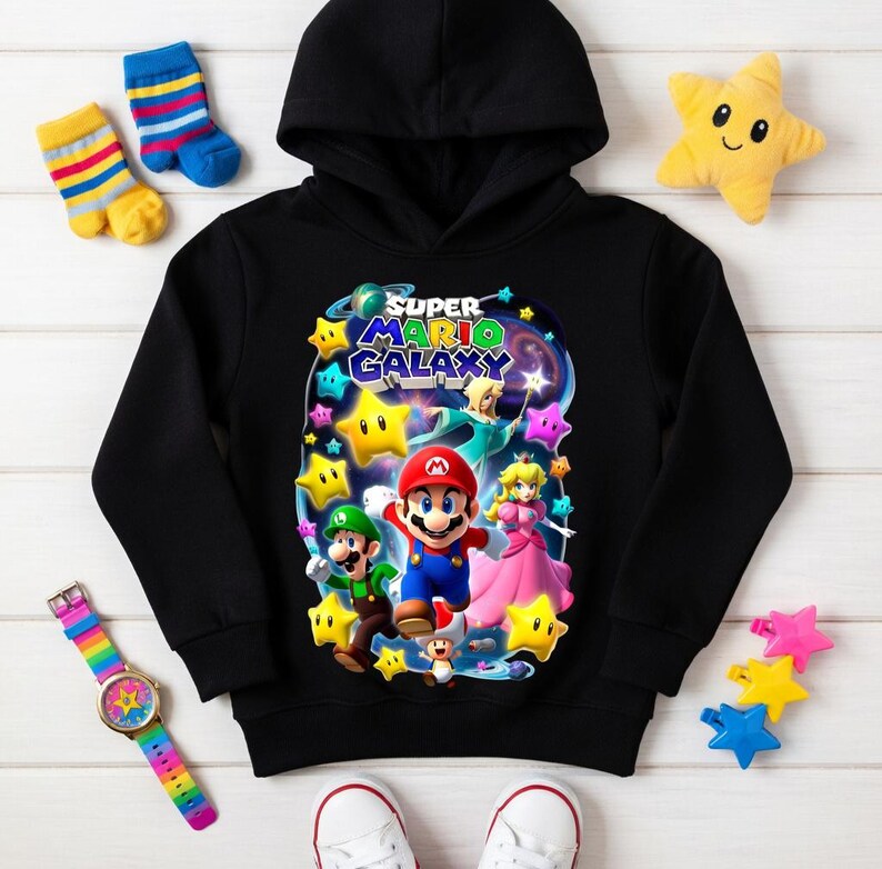 May include: Black hooded sweatshirt featuring a colorful graphic of Mario, Luigi, Princess Peach, and stars from the Super Mario Galaxy game. The sweatshirt has a drawstring hood and long sleeves. Additional items include a rainbow watch, star-shaped plush toys, and colorful socks.