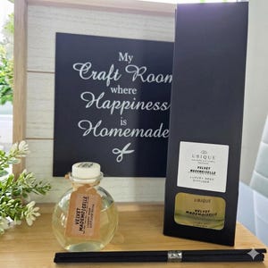 May include: A clear glass bottle of Velvet Mademoiselle luxury reed diffuser with a white cap and a brown paper tag, alongside a tall black rectangular box with a white label and a set of black reed sticks for home fragrance.