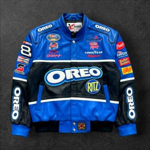 May include: Blue and black leather jacket with multiple patches and logos, including "OREO" and "RITZ". The jacket has a snap closure and a ribbed black bottom. The sleeves have "OREO" printed on them. The jacket is a racing style.