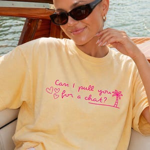 May include: A pale yellow short-sleeved t-shirt featuring the pink text "Can I pull you for a chat?" along with three small heart outlines and a palm tree graphic printed on the front centre of the garment.