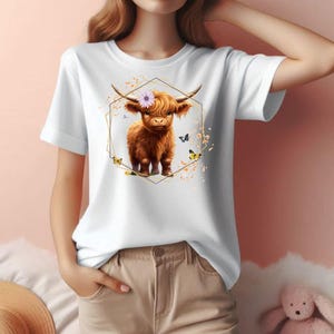 Pu&ograve; includere: T-shirt bianca con un'illustrazione di una mucca delle Highlands marrone con una corona di fiori, incorniciata da una forma geometrica dorata. Il design include farfalle ed elementi floreali. La maglietta ha maniche corte.