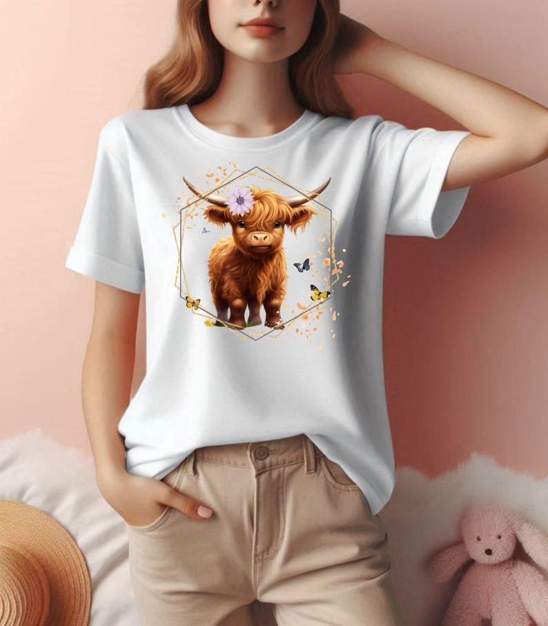 Pu&ograve; includere: T-shirt bianca con un'illustrazione di una mucca delle Highlands marrone con una corona di fiori, incorniciata da una forma geometrica dorata. Il design include farfalle ed elementi floreali. La maglietta ha maniche corte.