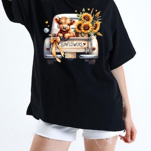 Pu&ograve; includere: T-shirt nera con una grafica di un cassone di camion vintage bianco pieno di girasoli e un vitello. Il cassone del camion ha la scritta "SUNFLOWERS" e una targa. Un fiocco giallo &egrave; legato al camion.