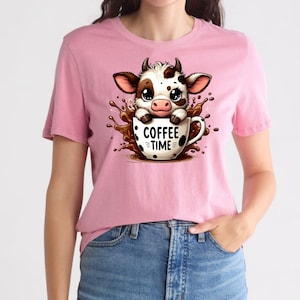 Pu&ograve; includere: T-shirt rosa con un disegno di mucca dei cartoni animati. La mucca, con macchie marroni e bianche, &egrave; in una tazza di caff&egrave; con la scritta "COFFEE TIME". Il disegno include uno schizzo di liquido marrone intorno alla tazza.