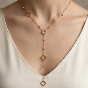 Peut inclure: Un collier en forme de Y de couleur or, orné de pendentifs en forme de trèfle et de petits accents de boules. Le collier est porté sur un haut blanc, créant un contraste qui met en valeur le design du bijou. C'est un bijou délicat.