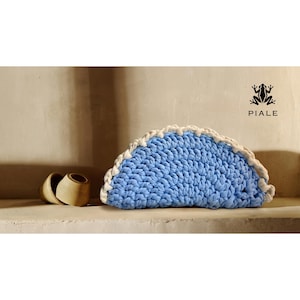 Op de afbeelding: Een lichtblauwe, halvemaanvormige gehaakte clutch met een witte rand. De clutch is gemaakt van dik garen en heeft een getextureerde uitstraling. De merknaam "PIALE" is zichtbaar.