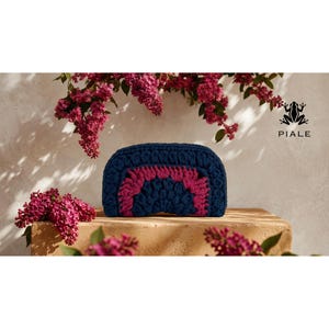 Op de afbeelding: Een marineblauwe gehaakte etui met een magenta regenboogontwerp. De etui wordt gepresenteerd op een bruin oppervlak, met roze bloemen op de achtergrond. De merknaam "PIALE" is rechtsboven zichtbaar.