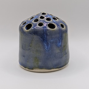 May include: A cylindrical ceramic flower frog with a domed top featuring multiple circular openings. The piece has a glossy glaze that transitions from a deep blue at the top to a muted, earthy green tone toward the unglazed, light-coloured base.