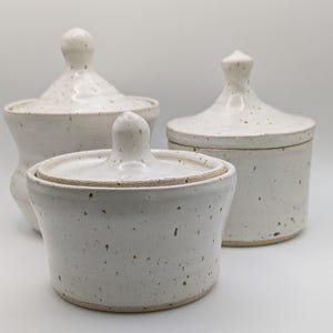 May include: Three ceramic lidded jars with a speckled white glaze. Each jar features a rounded body and a matching lid with a small, pointed knob handle. The vessels have a smooth, glossy finish with visible brown flecks distributed across the surface.