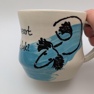 May include: A cream-coloured ceramic mug featuring a hand-painted light blue diagonal brushstroke. A black line drawing of a Saluki dog is centred over the blue paint. The text "heart" and "Saluki" is written in black ink on the left side.