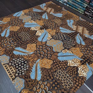 May include: A square fabric with a detailed batik pattern. The design features a mix of brown, black, and tan shapes, with accents of light blue feather-like elements. The fabric is folded, showcasing the intricate design.