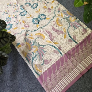 May include: Cream-colored fabric with a floral and bird pattern in teal, yellow, and purple. The fabric has a decorative border in a mauve color with an ornate design. The fabric is folded and displayed on a gray surface.