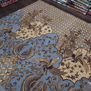 Könnte beinhalten: Ein detailliertes Textil mit blauem Hintergrund und aufwendigen braunen und goldenen Mustern. Das Design zeigt stilisierte Vögel, florale Motive und geometrische Formen. Der Stoff ist gefaltet, wodurch andere gemusterte Textilien sichtbar werden.