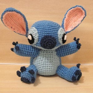 May include: A handmade crochet plush toy of a blue alien character with large ears, black eyes, and a dark blue nose. The plush has a light blue belly and dark blue accents on its feet and hands. The ears are pink inside.