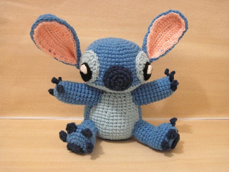 May include: A handmade crochet plush toy of a blue alien character with large ears, black eyes, and a dark blue nose. The plush has a light blue belly and dark blue accents on its feet and hands. The ears are pink inside.