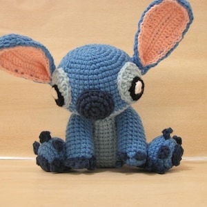 May include: A handmade blue crochet Stitch doll with large ears, grey accents, and black eyes. The plush toy is sitting with its legs outstretched. The inside of the ears are pink. The doll is made of yarn.
