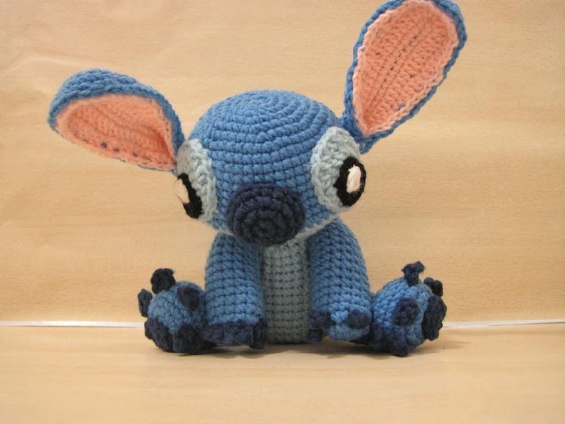 May include: A handmade blue crochet Stitch doll with large ears, grey accents, and black eyes. The plush toy is sitting with its legs outstretched. The inside of the ears are pink. The doll is made of yarn.