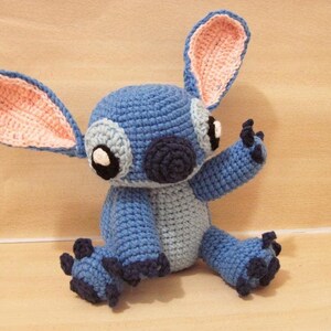 May include: A handmade blue crochet plush toy with large ears, black eyes, and a dark blue nose. The toy has a light blue belly and dark blue accents on its feet and hands. The ears have a pink interior.