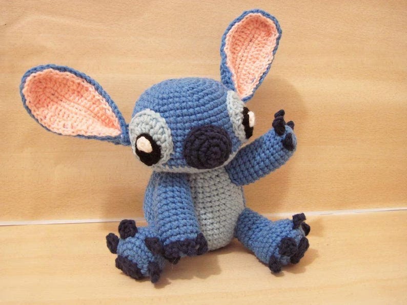 May include: A handmade blue crochet plush toy with large ears, black eyes, and a dark blue nose. The toy has a light blue belly and dark blue accents on its feet and hands. The ears have a pink interior.