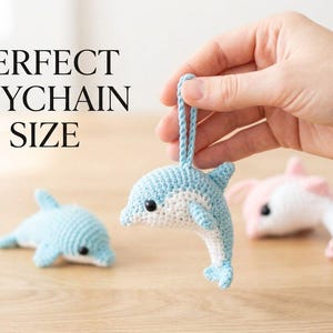 Puede incluir: Llaveros de delfines de ganchillo en azul claro, blanco y rosa. Un llavero es sostenido por una mano, mientras que otros dos descansan sobre una superficie de madera. El texto "PERFECT KEYCHAIN SIZE" se muestra en negro.
