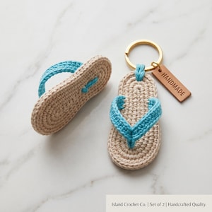 Può includere: Un set di due portachiavi a forma di infradito all'uncinetto con suole beige e cinturini blu brillante. Un'infradito è attaccata a un anello in metallo dorato con un'etichetta rettangolare in pelle marrone con la scritta HANDMADE.