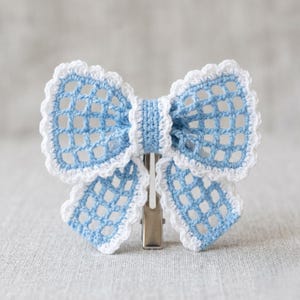 May include: A light blue and white crochet bow hair clip. The bow features a delicate open weave design with a scalloped edge. The clip is made of metal and is attached to the back of the bow. The bow is approximately 4 inches wide.