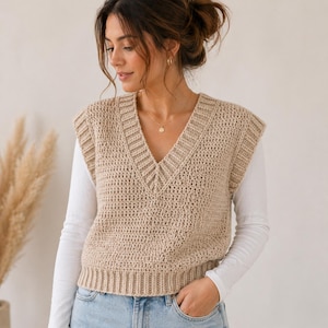 May include: A beige, hand-crocheted vest with a V-neck. The sleeveless garment has a ribbed hem and neckline. The vest is worn over a white long-sleeved shirt and blue jeans. The crochet stitch creates a textured appearance.