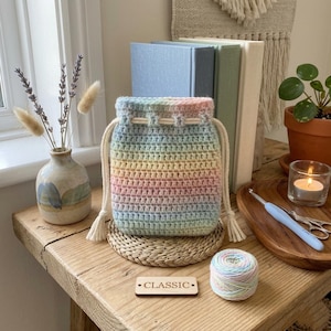 May include: A handmade, rainbow-colored crochet drawstring bag. The bag features a gradient of pastel colors and a cream-colored drawstring. A small wooden sign that says "CLASSIC" is next to the bag. A ball of yarn and a vase of flowers are also in the image.