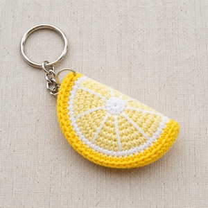 Può includere: Un portachiavi a forma di fetta di limone all'uncinetto. Il portachiavi presenta un design giallo brillante e bianco, con un bordo giallo e un portachiavi e catena in metallo argentato. La fetta di limone ha un centro bianco e linee bianche che si irradiano verso l'esterno.