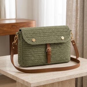 May include: A rectangular olive green crochet shoulder bag featuring a flap closure with two gold-tone metal snap buttons and a central brown leather strap with a gold-tone buckle. The bag includes a detachable brown leather shoulder strap.