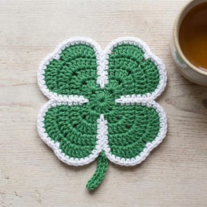 Può includere: Un sottobicchiere fatto a mano all'uncinetto a forma di quadrifoglio, verde e bianco. Il sottobicchiere è realizzato con filato verde con un bordo bianco e un gambo. È su una superficie di legno chiaro, con una tazza di tè sullo sfondo.