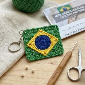 May include: A handmade green crochet keychain featuring the Brazilian flag design. The flag is square, with a blue circle in the center, surrounded by a yellow diamond and green border. A crochet hook, scissors, and a pattern are also visible.