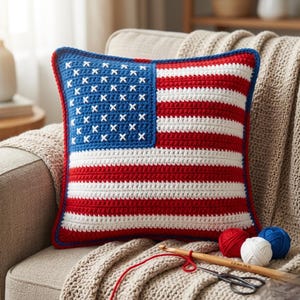 Può includere: Un cuscino all'uncinetto a forma di bandiera americana, con strisce rosse e bianche e un quadrato blu con stelle bianche. Il cuscino è su una poltrona beige, con gomitoli di lana rossi, bianchi e blu, un uncinetto e delle forbici.