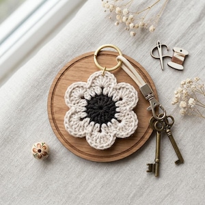 May include: A handmade crochet flower keychain with a black center and cream petals. The keychain has a gold ring and a cream-colored strap. A wooden coaster, vintage keys, and a decorative bead are also in the image.