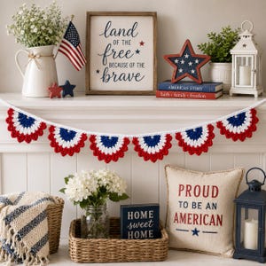 May include: A patriotic display featuring a crocheted red, white, and blue garland. The garland hangs above a shelf with a wooden sign that reads "land of the free because of the brave." Other decorations include a flag, star-shaped decor, and a pillow.